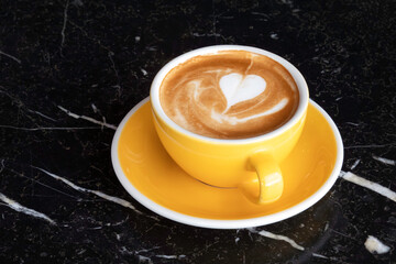 Cup of coffee latte in yellow ceramic mug, milk froth, heart shape on black table, fresh breakfast.