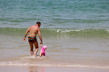Father leading child to the sea. Man with baby splashing in waves on a sandy beach, summer vacation concept
