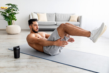 Shirtless young Arab man working out core muscles, doing abs exercises on yoga mat at home, full...
