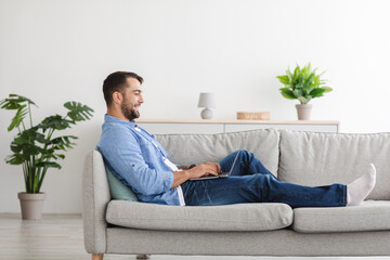Happy adult european man with beard sitting on sofa and typing on laptop in living room interior,...