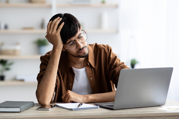 Exhausted indian man sitting in front of laptop, touching head