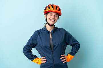 Young cyclist woman isolated on blue background posing with arms at hip and smiling - obrazy, fototapety, plakaty