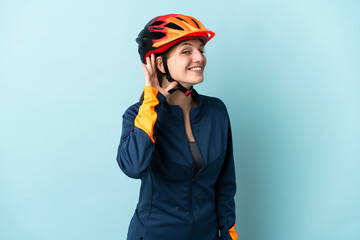 Young cyclist woman isolated on blue background listening to something by putting hand on the ear