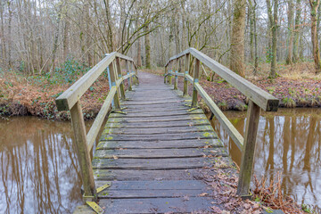 Obraz premium Small rustic wooden pedestrian bridge over the Leubeek river with reflection on the water surface, bare trees in the blurred background. Leudal Nature Reserve, Midden-Limburg in the Netherlands