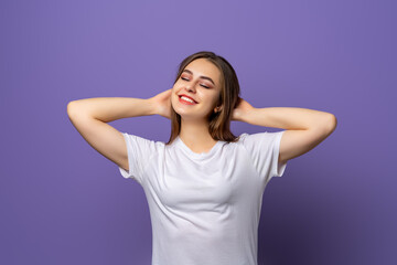 Portrait of dreaming brunette girl in blank white t shirt holding hands behind the had, close eyes and relax isolated on purple background