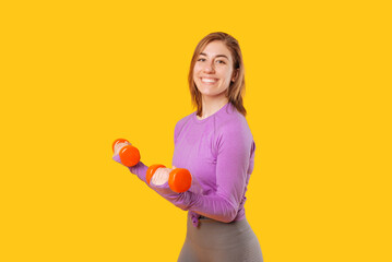Young smiling woman is lifting up some red dumbbells while smiling at the camera. Portrait over yellow background.