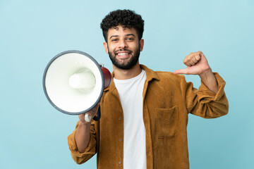 Young Moroccan man isolated on blue background holding a megaphone and proud and self-satisfied