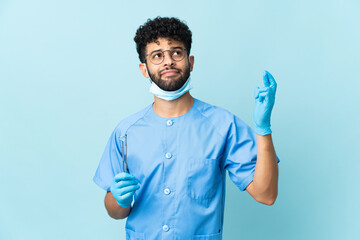Moroccan dentist man holding tools isolated on blue background with fingers crossing and wishing the best