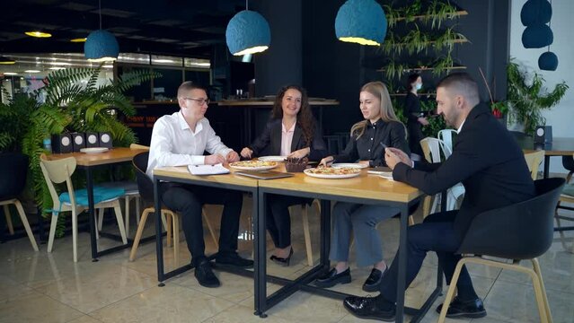 Teammates sitting in a buffet at lunchtime. Man and two women speaking and gesturing. Other man is concentrated on his smartphone.
