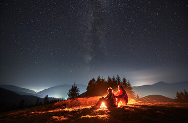 Side view of pair sitting one behind other on meadow near bright flame of bonfire and watching...