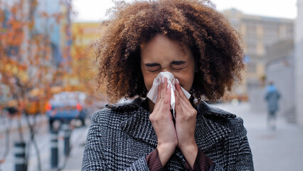 Sick ill african american woman with curly hair blowing runny nose into tissue paper outdoors cold...