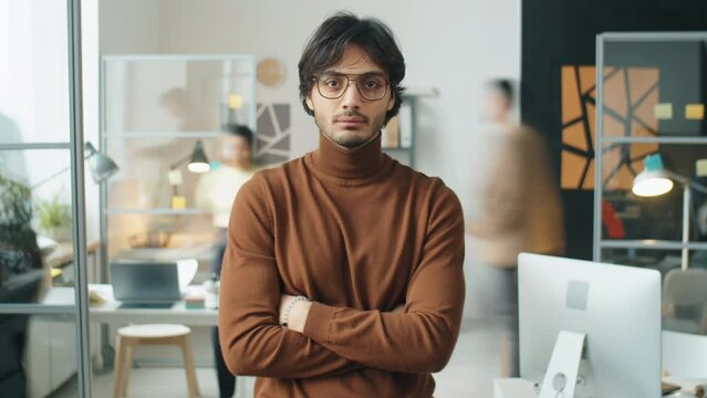 Time lapse shot of handsome middle eastern businessman in glasses and brown turtleneck standing with arms crossed in office and posing on camera during workday
