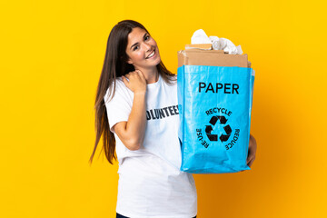 Young brazilian girl holding a recycling bag full of paper to recycle isolated on yellow background laughing