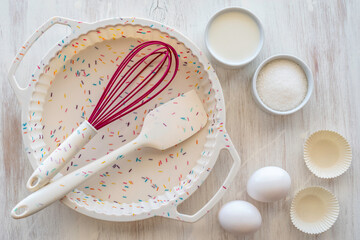 Silicone cake form, spatula and whisk on white wooden background.
