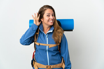 Middle age mountaineer woman with a big backpack over isolated background listening to something by putting hand on the ear