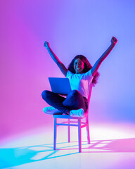 Excited young black lady sitting on chair with laptop, raising arms up, celebrating success in neon light
