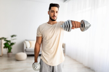 Portrait of handsome young Arab guy exercising with dumbbells at home during covid-19 lockdown, selective focus