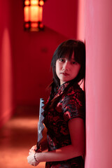 A chinese girl is standing against a red wall in a Chinese dress qipao looking at the camera seriously