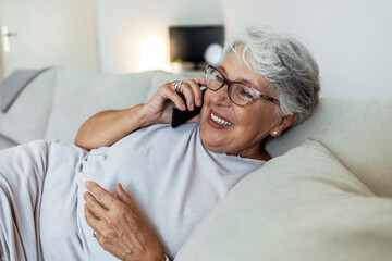 Obraz premium Close up of a senior woman using a phone in the living room. Smiling senior woman using phone sitting on couch at home. Cheerful old lady talking on cell phone and smiling.