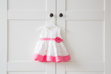 White pink new baby dress on hanger on door handle of wardrobe at home. Closeup. Front view. Clothes preparing for party, holidays or other celebrations.
