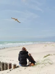 Selbstklebende Fototapeten Junge Frau sitzt und umarmt ihren Hund am Strand bei Sonnenschein, eine Möwe fliegt über den beiden © SINNBILD Design