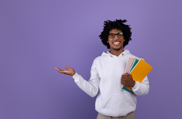 Education concept. Positive black teen guy with notebooks offering empty space for your ad on violet studio background