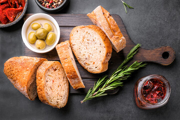Slices of Ciabatta bread  on wooden board with olives, dried tomatoes, oil, rosemary and herbs on rustic wooden background. top view.