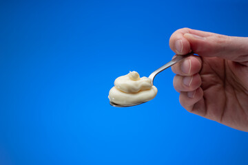 Fresh homemade mayonnaise in a small spoon held between male fingers. Close up studio shot, isolated on blue background