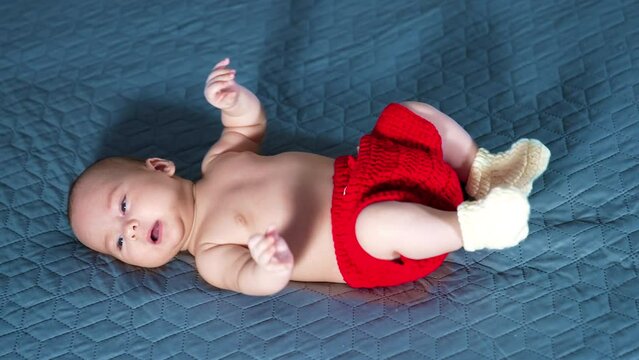 Active toddler boy in red shorts and white socks. Caucasian baby moves feet cheerfully while lying on the bed. Grey backdrop.