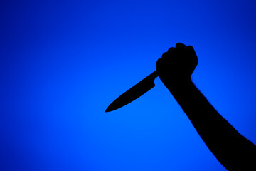 Silhouette on a male arm holding a long kitchen knife threateningly. Close up studio shot, isolated on blue background