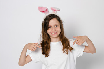 Cute dark-haired girl 9 years old. With bunny ears on the head. Pointing at a white T-shirt. Easter Bunny. close-up. High quality photo. copy space 