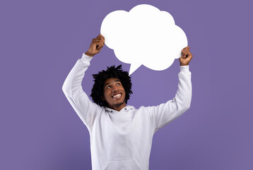 Handsome black teenager holding empty speech bubble above his head on violet studio background,...