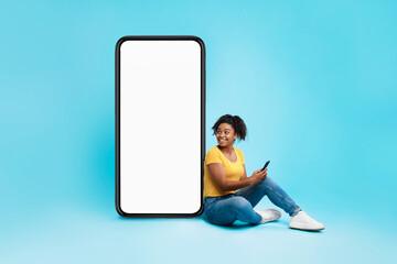 Cheerful black woman sitting near huge smartphone with empty screen, holding mobile device on blue background, mockup