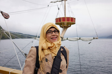 Portrait of smiling muslim woman on board Hakone sightseeing cruise ship at Lake Ashi with mountain fog and cloudy sky background. Happy expression.