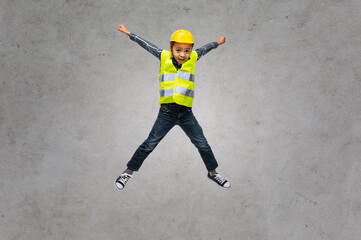 Fototapeta na wymiar building, construction and profession concept - smiling little boy in yellow safety vest and helmet jumping over grey concrete background