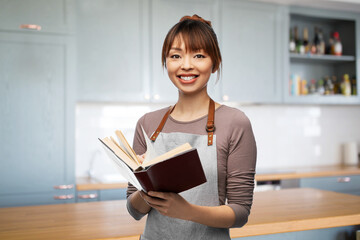 Fototapeta premium cooking, culinary and people concept - happy smiling woman in apron with open cook book over home kitchen background