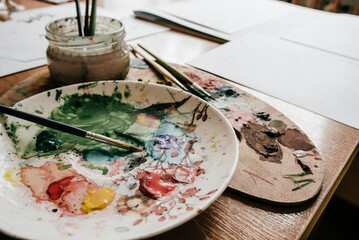 Artist workplace, watercolor paints, paint brushes on a palette, dirty papers, wood table. Low...