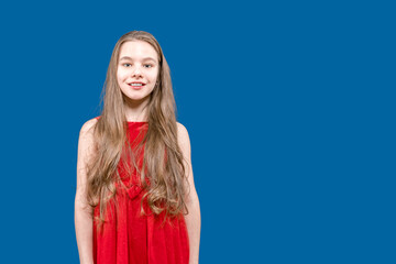 Fototapeta premium A cute teenage girl with long blond hair is standing on a blue background smiling. Pupil.
