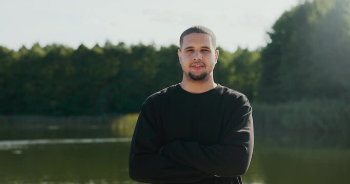 A Man In A Black Sweatshirt Is Standing In Background Of A Lake, He Has His Arms Crossed Over Chest, He Is Looking Into The Camera With A Serious Face, A Handsome Guy Is Spending Time Outdoors