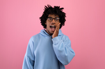 Cool black teen in glasses opening mouth in shock, shouting WOW, touching cheek, looking at camera on pink background
