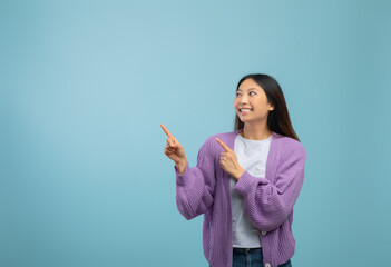 Cool asian lady pointing and looking at free space over blue studio background, showing exciting...
