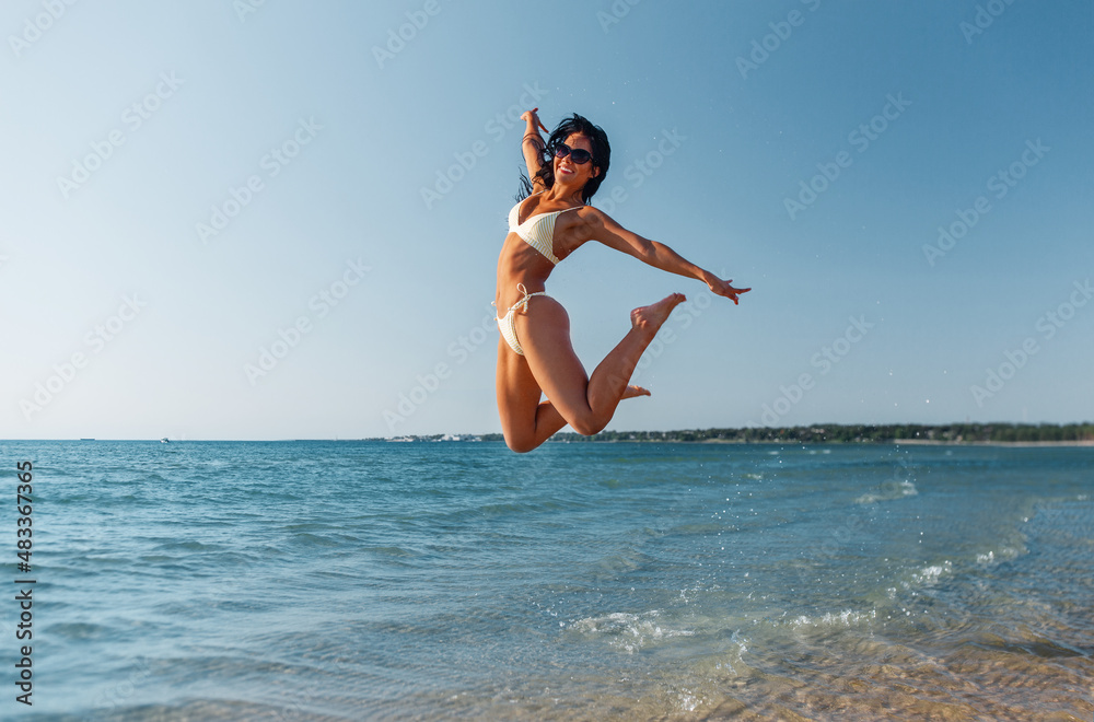 Wall mural people, summer and swimwear concept - happy smiling young woman in bikini swimsuit jumping on beach