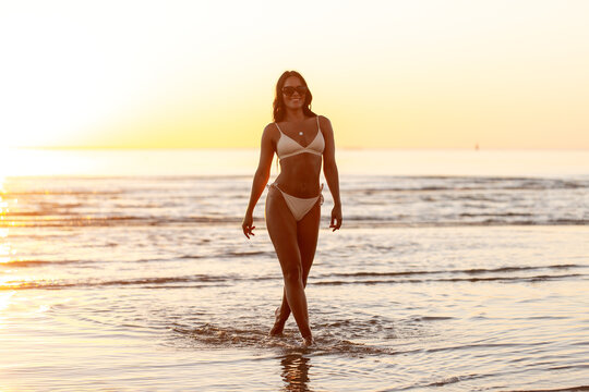 people, summer and leisure concept - happy smiling young woman in bikini swimsuit on beach over sunset