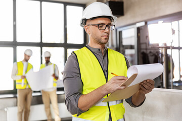 architecture, construction business and building concept - male architect in helmet and safety west with clipboard working at office