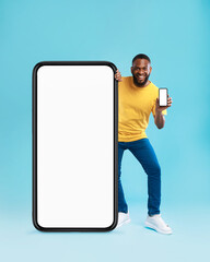 Handsome black guy standing near giant cellphone with blank screen, showing mobile device on blue background, mockup