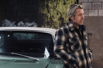 Blond man in checkered jacket leans against the drivers side of a classic american muscle car.