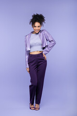 full length of cheerful african american woman in violet leather jacket and trousers posing with hand on hip on purple.