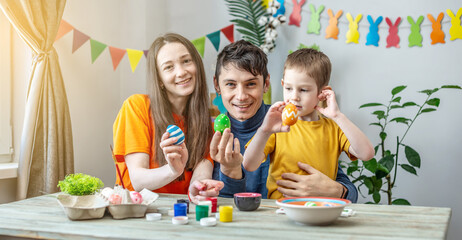 Mom, dad and little son in bright clothes is coloring eggs in a decorated room. Concept of family preparation for Easter, festive spring mood