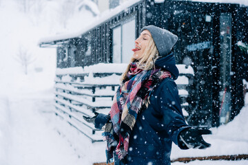 A young woman in a winter jacket, hat and scarf sticks out her tongue to catch snowflakes during a snow storm.