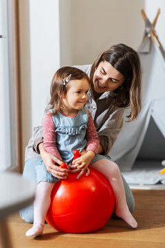 family, leisure and people concept - happy mother playing with little baby daughter bouncing on hopper ball at home